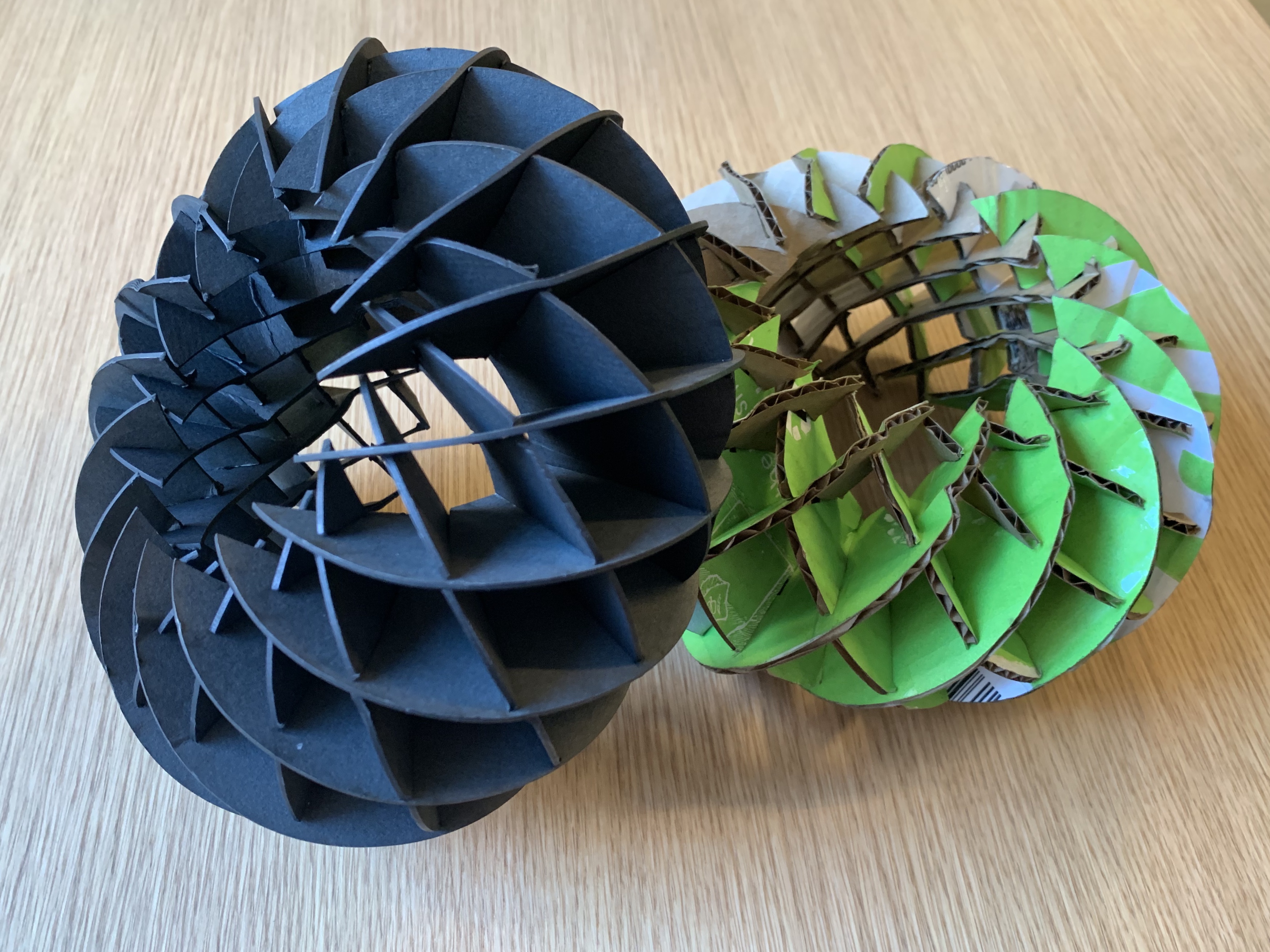 two round sculptures made of cardboard pieces laser cut into moon shapes with slots in them. One is black, one is green and brown.